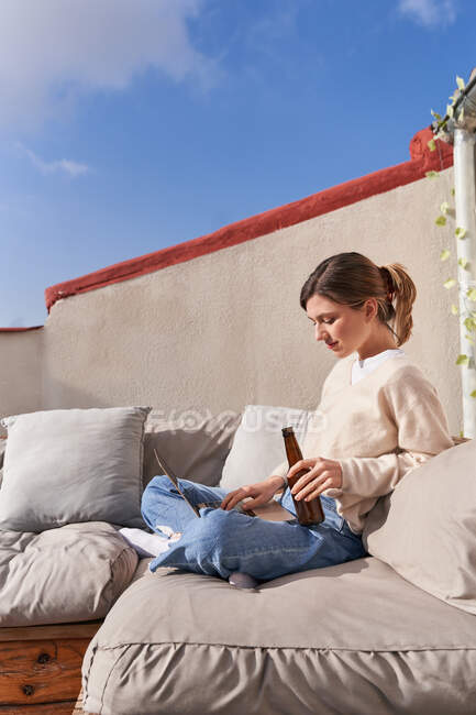 Vista lateral de una joven hembra sentada con las piernas cruzadas tomando una botella de cerveza y usando un netbook moderno en un cómodo sofá en el balcón en un día soleado - foto de stock
