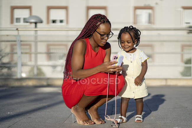 Calm African American woman with braids in red outfit squatting down and showing smartphone to little daughter on street against residential building in sunlight — Stock Photo
