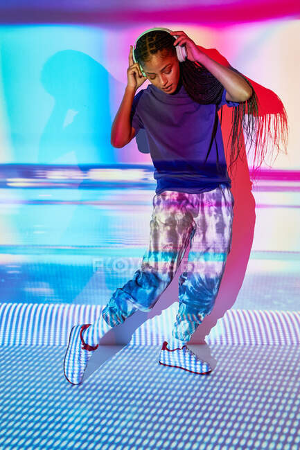 Vue latérale du jeune millénium féminin dominicain à la mode avec de longues tresses afro debout sur le sol et regardant vers le bas tout en écoutant de la musique dans les écouteurs dans la chambre avec un éclairage géométrique coloré — Photo de stock