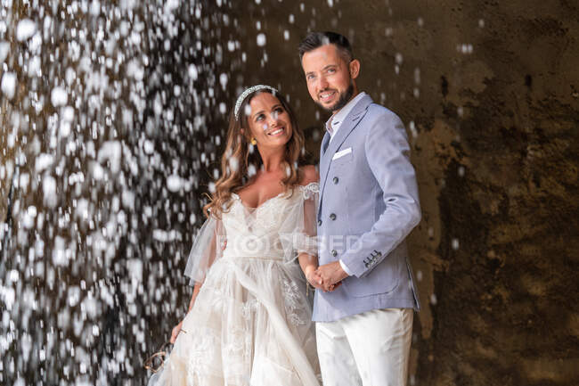 Позитивна весільна пара в елегантному вбранні, що стоїть на валуні біля швидкого каскаду під час весільного торжества в природі — стокове фото