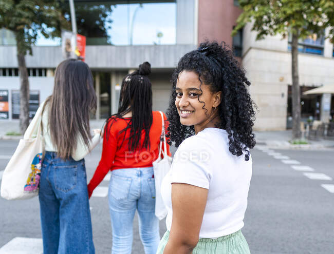 Femme afro-américaine souriante avec des cheveux bouclés dans des vêtements décontractés debout et regardant par-dessus l'épaule à la caméra tout en marchant avec des copines sur la rue de la ville en plein jour — Photo de stock