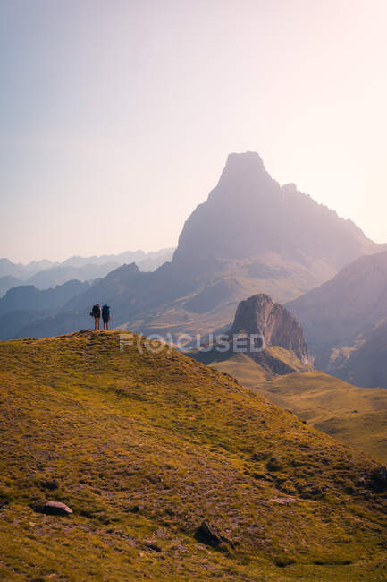 Далекие анонимные туристы, стоящие на травянистой вершине холма, любуясь грубым горным хребтом против безоблачного неба в природе Испании — стоковое фото