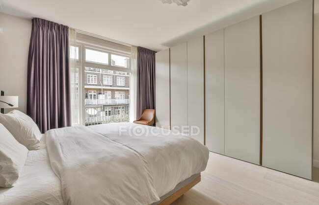 Cama confortável e armário de estilo minimalista localizado perto da janela com cortinas no quarto moderno — Fotografia de Stock