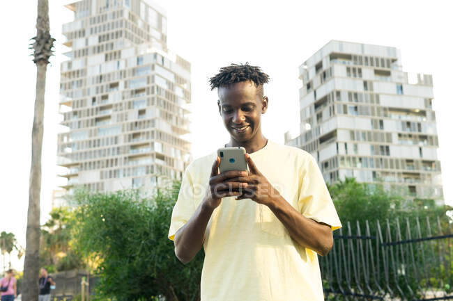 Улыбающийся афроамериканец в обычной футболке отправляет смс на мобильный телефон и смотрит на экран города с ладонями — стоковое фото