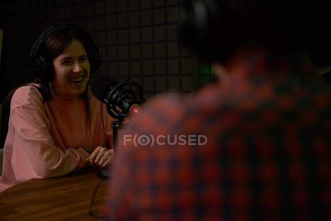 Sorridi giovane conduttrice radiofonica femminile con cuffie sedute a tavola con microfono e comunicanti con collega anonimo durante la registrazione del podcast in studio — Foto stock