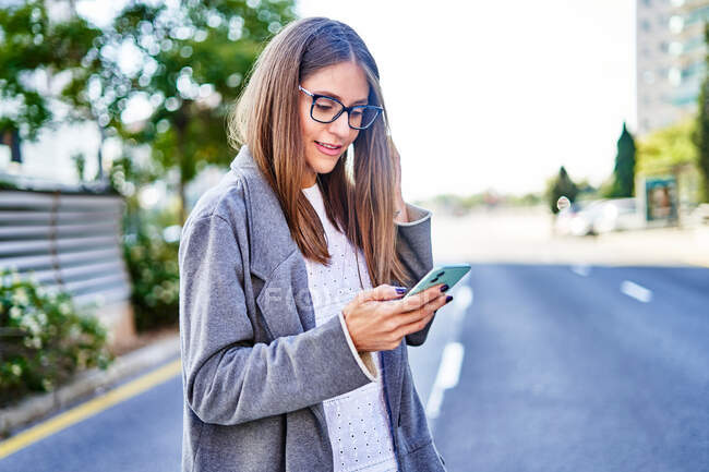Позитивна жінка в офіційному носінні використовує мобільний телефон під час ходьби по міській вулиці для роботи — стокове фото