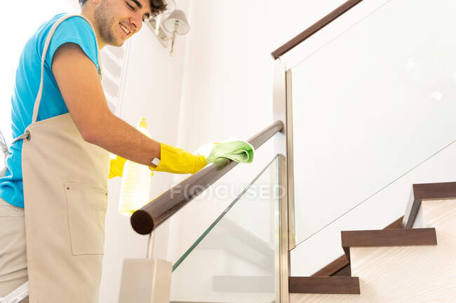 Vista lateral de la cosecha feliz empleado masculino en uniforme y guantes de goma sonriendo mientras se limpia el polvo de la barandilla de pie en la escalera durante el trabajo en la casa moderna - foto de stock