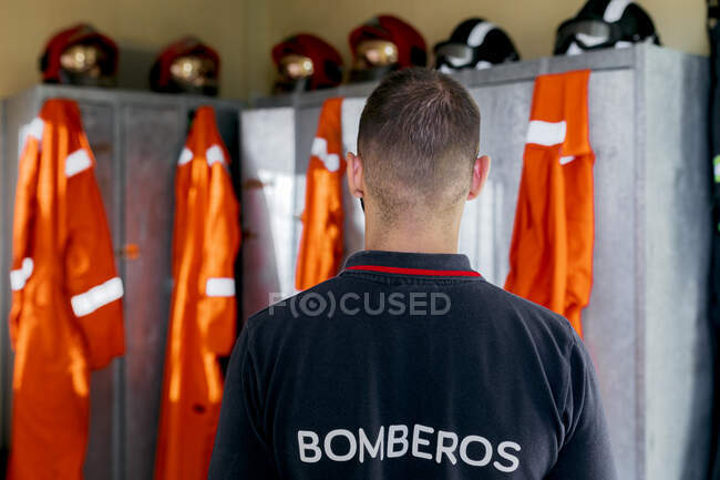 Pompier avec le dos tourné dans les vestiaires près d'une rangée de casiers en métal avec des casques sur le dessus et uniforme orange accroché — Photo de stock