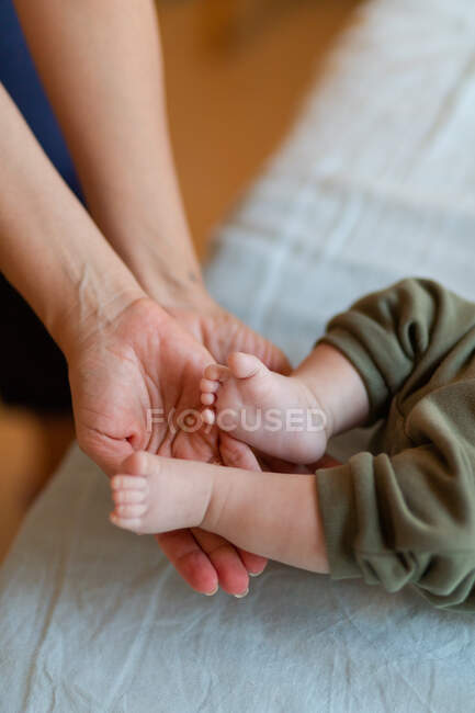 From above of crop anonymous mother massaging small feet of adorable barefoot baby lying on bed in sunny morning — Stock Photo