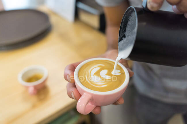 Anonimo barista etnico versando latte in tazza con caffè mentre in piedi al bancone con caffettiera e stoviglie in caffetteria — Foto stock
