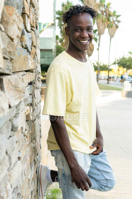 Hombre joven afroamericano positivo con sonrisa dentada usando ropa casual de pie y inclinada doblada en la pierna de rodilla en la pared de piedra - foto de stock