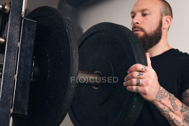 Мощный спортсмен в спортивной форме стоит с черной пластиной в руках и смотрит в сторону во время тренировки в тренажерном зале — стоковое фото