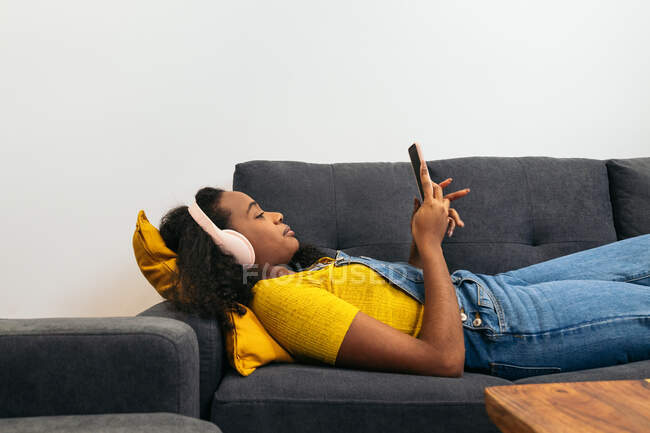 Young African American female with curly hair in headphones and casual clothes lying on comfortable gray sofa and browsing on smartphone in light room at home — Stock Photo