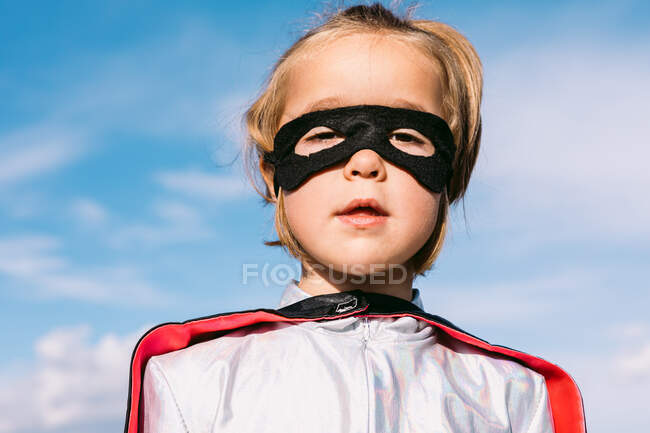 Lindo niño usando disfraz de superhéroe y máscara de ojos de pie contra el cielo azul y mirando a la cámara - foto de stock