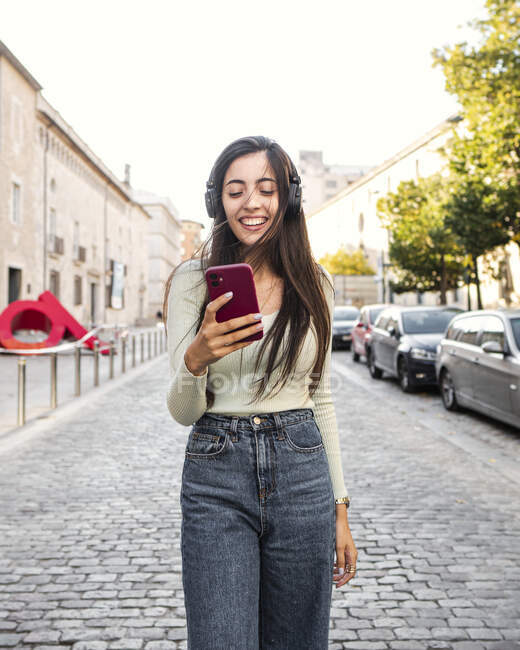 Fröhliche junge Frau mit langen braunen Haaren in lässiger Kleidung, die bei Tageslicht Musik über Kopfhörer auf der Straße hört — Stockfoto
