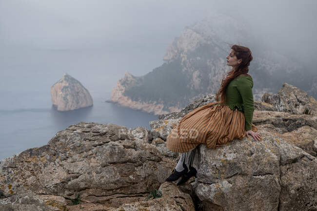 Visão lateral de corpo inteiro de mulher sonhadora em roupas antiquadas sentadas na borda de penhasco pedregoso perto do mar em tempo nebuloso — Fotografia de Stock