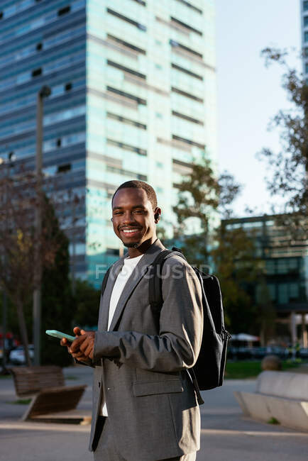Sonriente hombre de negocios afroamericano en ropa formal con mochila navegando por el teléfono móvil en el centro de la ciudad y mirando a la cámara con sonrisa dentada - foto de stock
