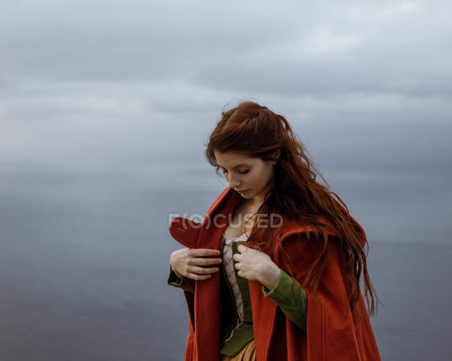 Dreamy female with long hair in Victorian styled outfit with cape standing above endless sea against cloudy sky in nature — Stock Photo