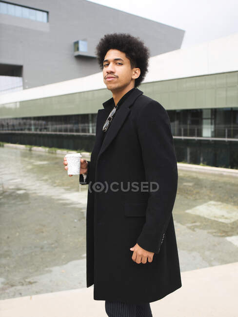 Side view of self assured young ethnic male millennial with dark Afro hair in trendy outfit standing near fountain with cup of takeaway coffee and looking at camera — Stock Photo