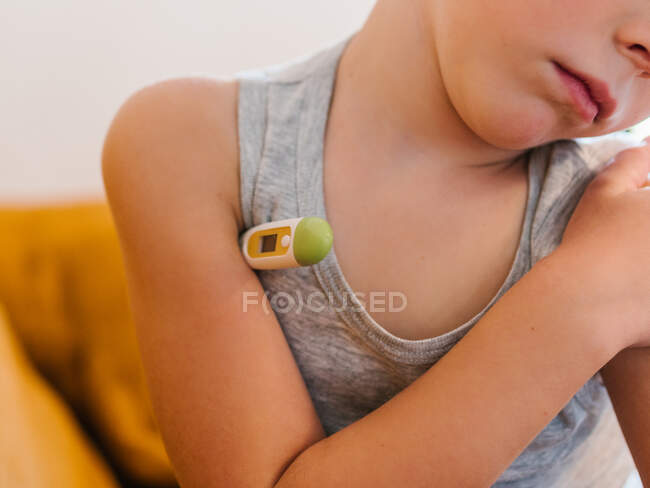 Niño enfermo anónimo midiendo la temperatura con termómetro electrónico mientras está sentado en el sofá en casa y tiene gripe - foto de stock