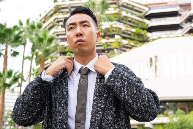 Ben vestito giovane imprenditore maschio asiatico in cravatta guardando lontano mentre passeggiava sulla strada contro edifici moderni in città — Foto stock