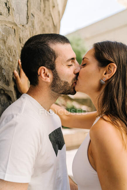 Vista lateral de la joven pareja étnica enamorada en ropa casual abrazándose y besándose de pie cerca de la pared de piedra en la calle de la ciudad - foto de stock