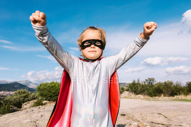 Menina pequena em traje de super-herói levantando punhos estendidos para mostrar poder enquanto estava de pé contra o céu azul claro — Fotografia de Stock