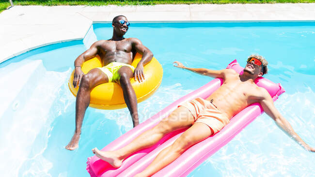 Mehrrassige männliche Freunde in Badehosen liegen auf aufblasbarem Ring und Matratze, während sie sich an sonnigen Sommertagen im Pool sonnen — Stockfoto