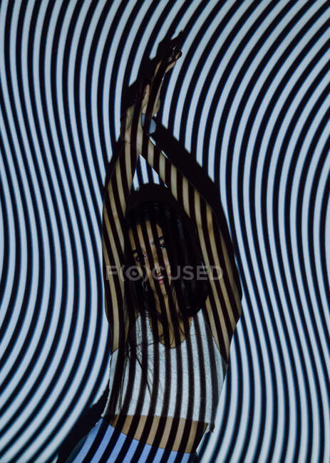 Модна етнічна жінка тисячоліття з піднятими руками і темним волоссям дивиться на камеру в хвилястих смугах від проектора світла — стокове фото