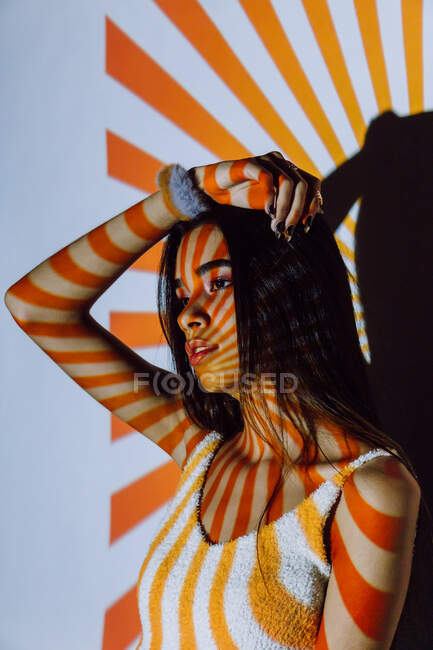 Fresco giovane donna etnica in crop top con strisce sul corpo dalla luce del proiettore guardando altrove — Foto stock