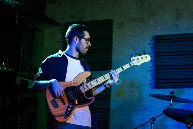 Серьезный молодой человек, играющий на бас-гитаре во время выступления в легком клубе с неоновой подсветкой — стоковое фото