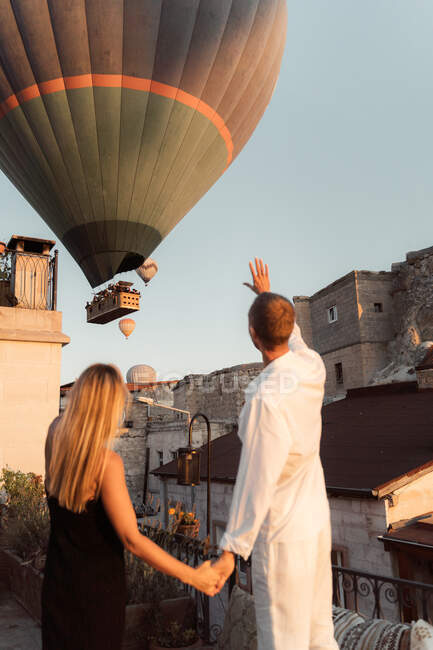 Rückansicht eines bis zur Unkenntlichkeit liebenden Mannes, der die Hände einer Frau hält und mit Heißluftballons am Abendhimmel in Kappadokien auf die Dachterrasse zeigt — Stockfoto