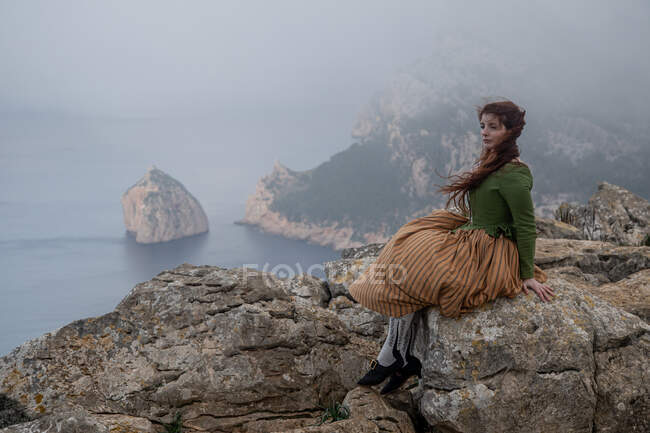 Повний вид збоку тіла мрійливої жінки в старомодному одязі, що сидить на краю кам'яної скелі біля моря в туманній погоді — стокове фото