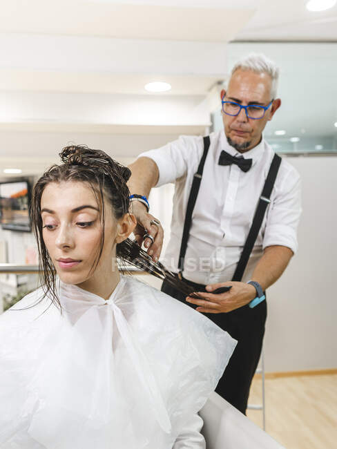 Клиентка в белом плаще смотрит в сторону, в то время как парикмахер работает с волосами — стоковое фото