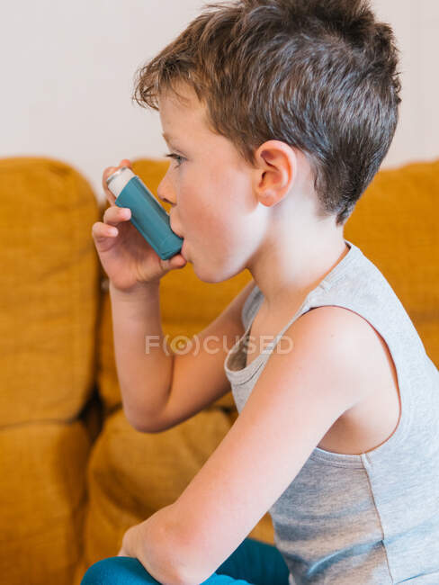 Seitenansicht eines kleinen Jungen mit Asthma beim Inhalieren, während er zu Hause auf dem Sofa sitzt — Stockfoto