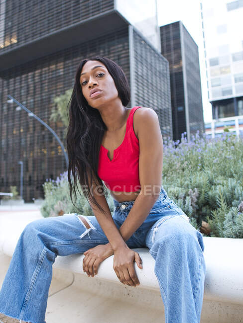 Selbstbewusste Afroamerikanerin in Top und Jeans blickt in die Kamera, während sie auf einer Blumenhöhle mit grünen Pflanzen auf der Straße mit modernen mehrstöckigen Gebäuden sitzt. — Stockfoto