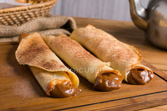 Fresh rolled crepes filled with sweet dulce de leche filling served on wooden cutting board on table with kettle in kitchen — Stock Photo