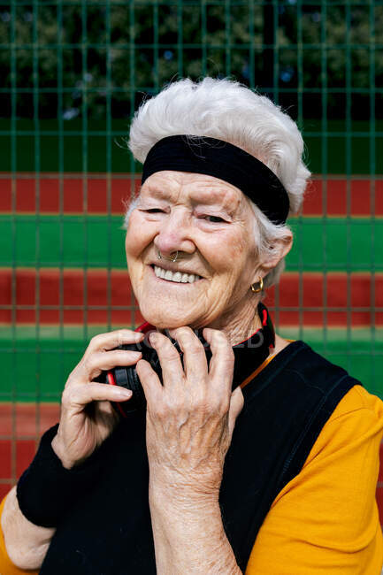 Позитивная зрелая женщина с проколотым носом и наушниками в спортивной одежде, смотрящая в камеру во время тренировки на спортивной площадке — стоковое фото
