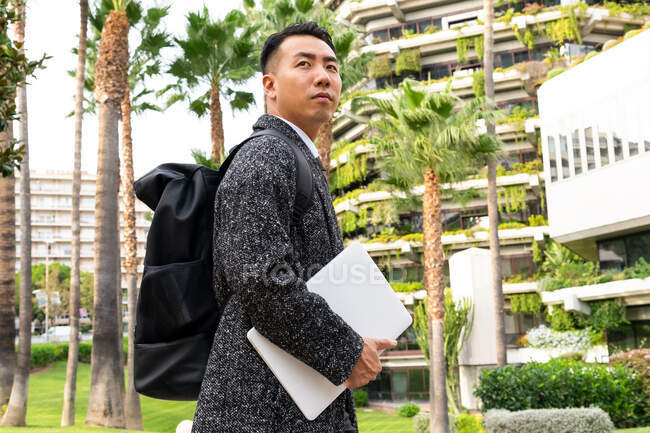 Side view of contemplative young ethnic male entrepreneur with rucksack and netbook looking away against modern urban buildings and palm trees — Stock Photo