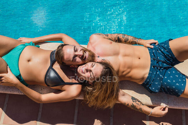 From above of delighted romantic couple in swimsuits lying together on border of swimming pool on sunny resort during vacation — Stock Photo