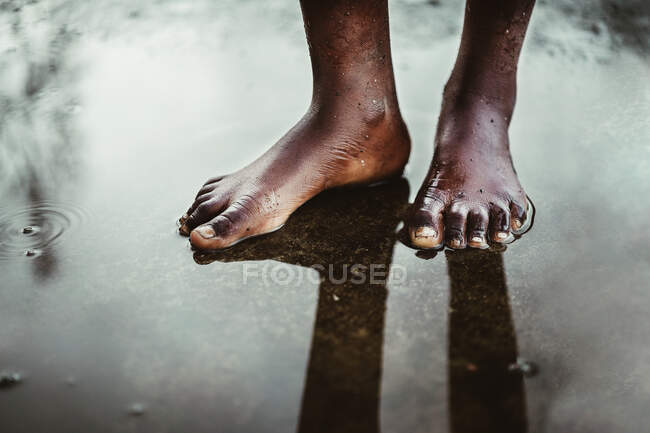 Сверху на урожай неузнаваемый черный босоногий ребенок, стоящий в маленькой луже на асфальтированной дороге на улице на острове Так Том и Прнсипи при дневном свете — стоковое фото