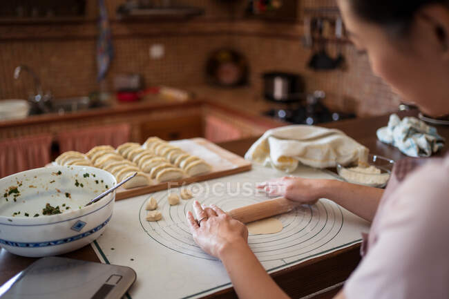 Женщина в фартуке катит тесто на столе, готовя пельмени с мясом на кухне — стоковое фото