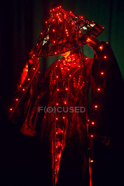 Mulher enigmática irreconhecível em roupa tradicional criativa e headwear vietnamita com iluminação vermelha em pé no estúdio escuro em fundo preto durante a performance — Fotografia de Stock