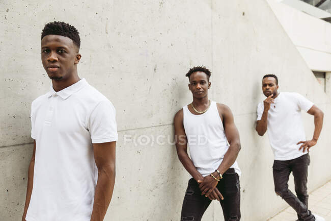 Cool African American male friends wearing trendy clothes with white t-shirt standing near building and looking at camera — Stock Photo
