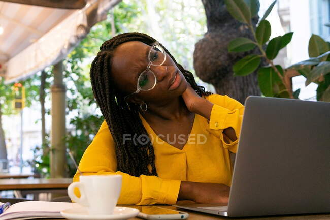 Thoughtful African American female freelancer in eyeglasses looking away while sitting at table with netbook and cup of hot drink in cafeteria — Stock Photo