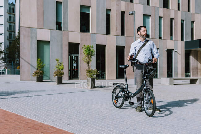 Longitud completa del trabajador ejecutivo en ropa formal de pie con bicicleta cerca del moderno edificio de oficinas - foto de stock
