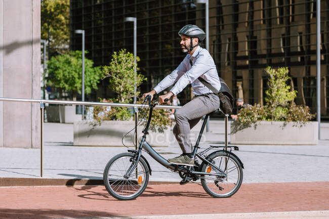 Vista lateral de longitud completa del trabajador masculino barbudo en casco en carretera lateral con bicicleta en camino al trabajo - foto de stock
