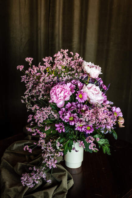 Ramo de peonías frescas y coloridas y crisantemos en jarrón blanco colocado sobre mesa de madera en habitación oscura - foto de stock