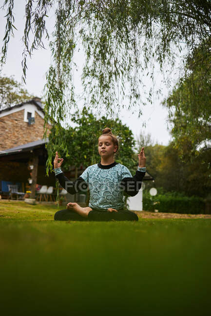 Full body of tranquil barefoot girl with closed eyes sitting on grassy lawn in Padmasana posture on grassy courtyard against building — Stock Photo