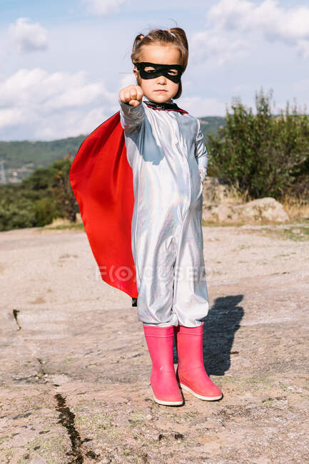 Полное тело маленькой девочки в костюме супергероя, поднимающей протянутые кулаки за демонстрацию силы, стоя на скалистом холме — стоковое фото